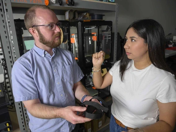 Matthew Briggs talks with Ruth Salazar while holding an adaptive foot pedal in a lab with 3D printers in the background