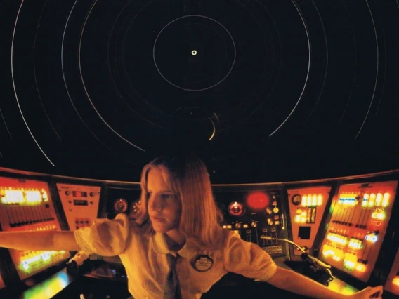 A planetarium operator sits at a large circular console filled with glowing buttons and switches, surrounded by projected star trails on the dome above.