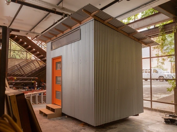 an 8-by-12-foot shelter with metal siding and an orange door sitting inside a large manufacturing shop