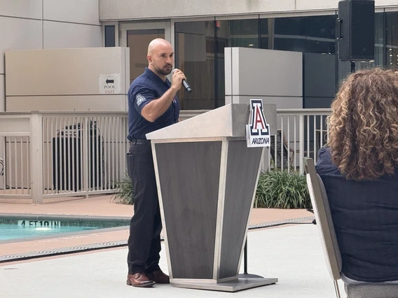 A man in a navy blue shirt speaking into a microphone at a podium. The podium is positioned near a swimming pool and features the University of Arizona logo with the word "ARIZONA" on it. In the background, there is a railing and plants adjacent to a modern building with large windows.