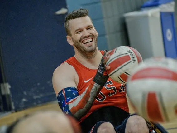 A man holding a rugby ball while wearing a red Team USA jersey with a sleeve on his arm with an American flag design smiles during a practice session. The gym environment is visible in the background.