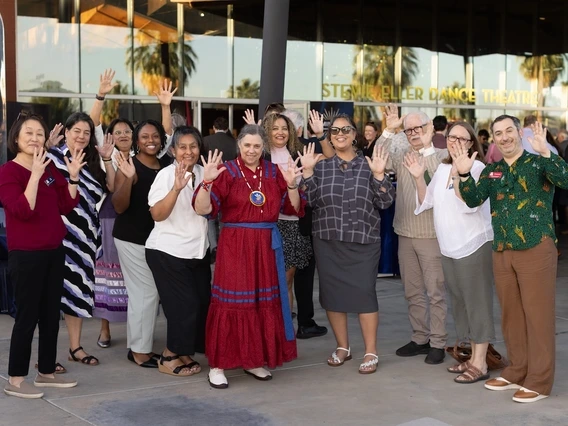 A group of people stand outside the Stevie Eller Dance Theatre, smiling and waving toward the camera. The group includes men and women in a variety of professional and traditional attire, gathered in front of the building’s glass entrance during a reception.