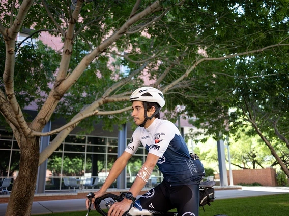A cyclist wearing a white helmet and blue jersey rides a bike on a tree-lined path next to a building with large windows.