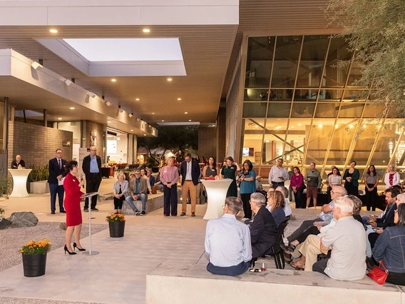 A woman speaks at a podium to an audience during an outdoor evening event at a modern university building.