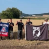 Five people stand in a golden wheat field holding two flags—one red, white, and blue Arizona Wildcats flag and one black POW*MIA flag with the words “You Are Not Forgotten.” They wear casual outdoor clothing, some with boots and hats, and make the University of Arizona “Wildcat” hand sign against a backdrop of rolling farmland and blue sky.
